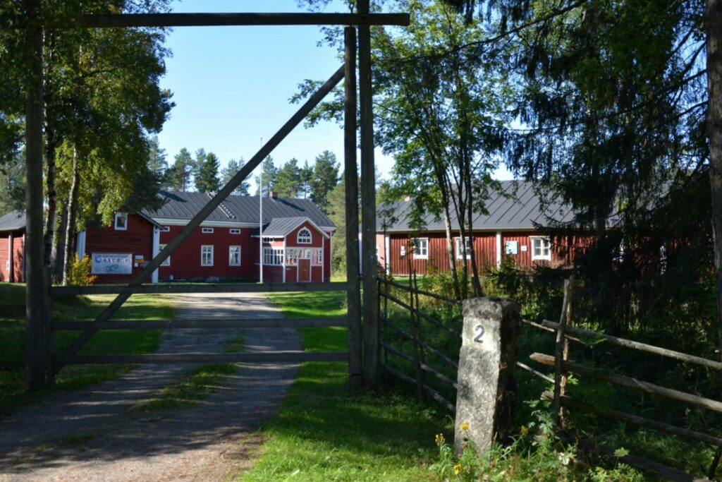 Punaisia vanhoja rakennuksia metsäisessä maastossa kesllä. Etualalla puinen portti ja kivinen kilometripylväs