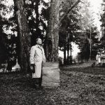 Mies seisoo mustavalkoisessa kuvassa kivisen muistimerkin vieressä, taustalla puita.