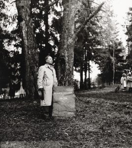 Mies seisoo mustavalkoisessa kuvassa kivisen muistimerkin vieressä, taustalla puita. 