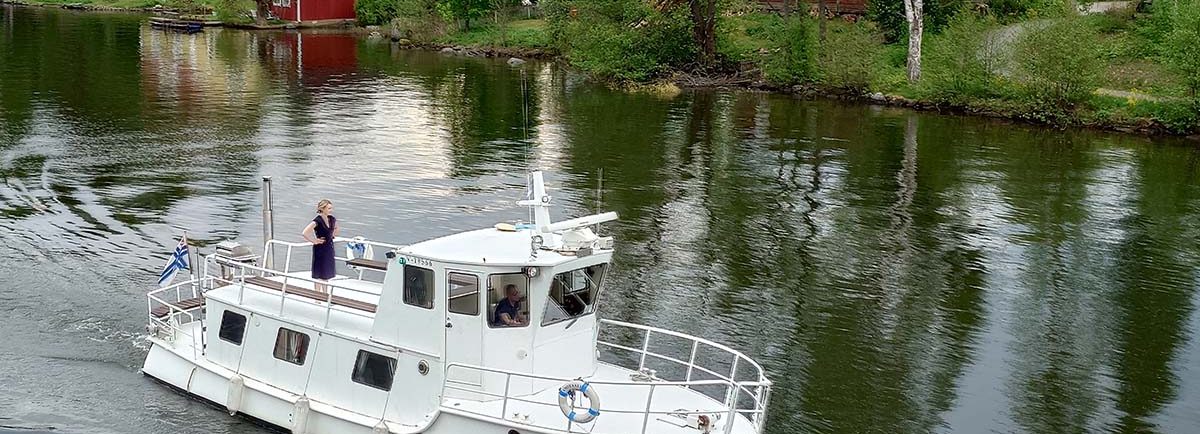 Valkeakosken vesistöistä kaupunkimaisemaa. Iso moottorivene, jonka kannella seisoo nainen ja nauttii maisemasta. Taustalla on idyllisiä, vanhoja puurakennuksia.