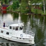 Valkeakosken vesistöistä kaupunkimaisemaa. Iso moottorivene, jonka kannella seisoo nainen ja nauttii maisemasta. Taustalla on idyllisiä, vanhoja puurakennuksia.