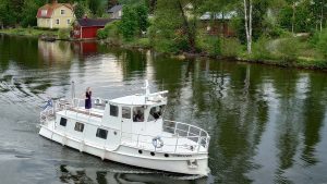 Valkeakosken vesistöistä kaupunkimaisemaa. Iso moottorivene, jonka kannella seisoo nainen ja nauttii maisemasta. Taustalla on idyllisiä, vanhoja puurakennuksia.