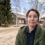 nainen etualalla ja vanha harmaa rakennus sekä puita takana