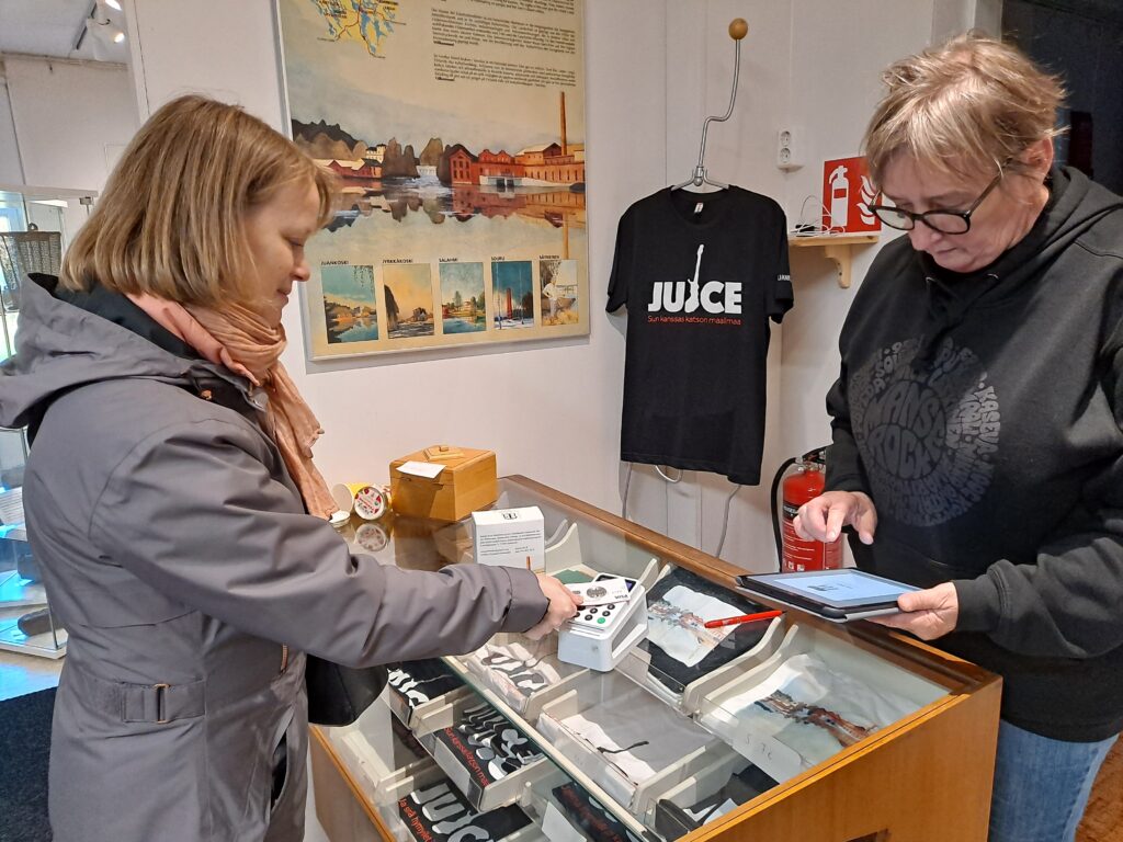 Nainen maksaa pankkikortilla museon kassalla. Oikealla nainen käyttää tablettia. Kassapöydällä ja taustalla museokaupan tuotteita.