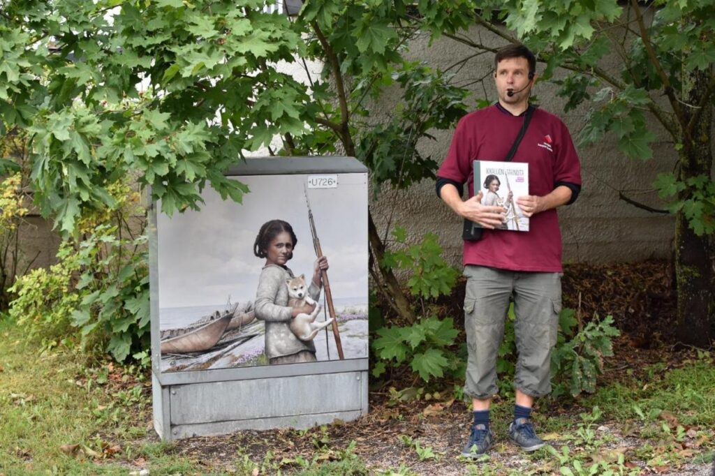 Mies seisoo sähkökaapin vieressä. Sähkökaappiin on maalattu esihistoria-aiheinen kuva tytöstä ja koirasta. Taustalla muutama puu ja seinää.