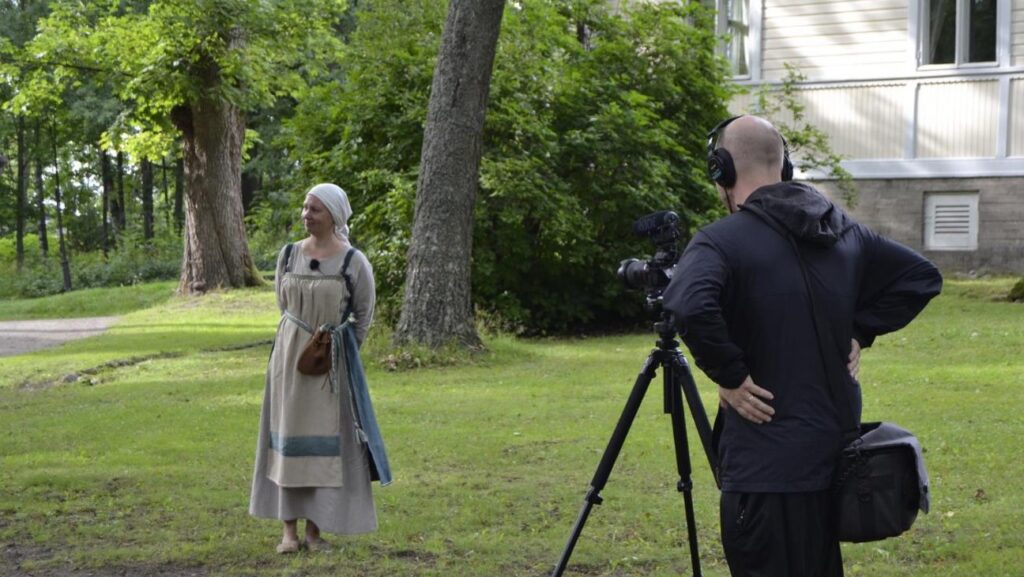 Nainen seisoo historiaa elävöittävässä asussa vasemmalla. Oikealla videokuvaaja selin. Taustalla vanha rakennus ja puita.