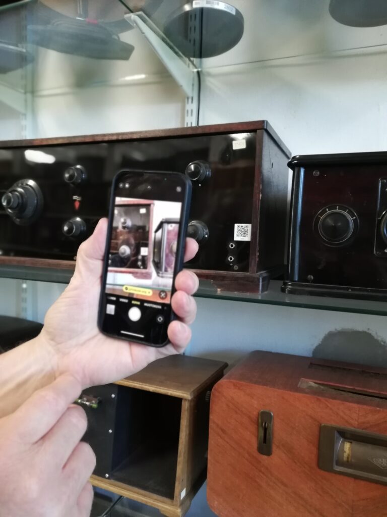 Kädessä puhelin, jossa näkyy vanhoja radioita. Taustalla samat vanhat radiot hyllyllä.