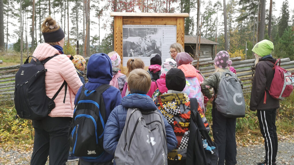 Koululaisia ja kaksi aikuista ulkona museon opastaulun edessä. Takana riukuaita, metsää ja harmaa rakennus.