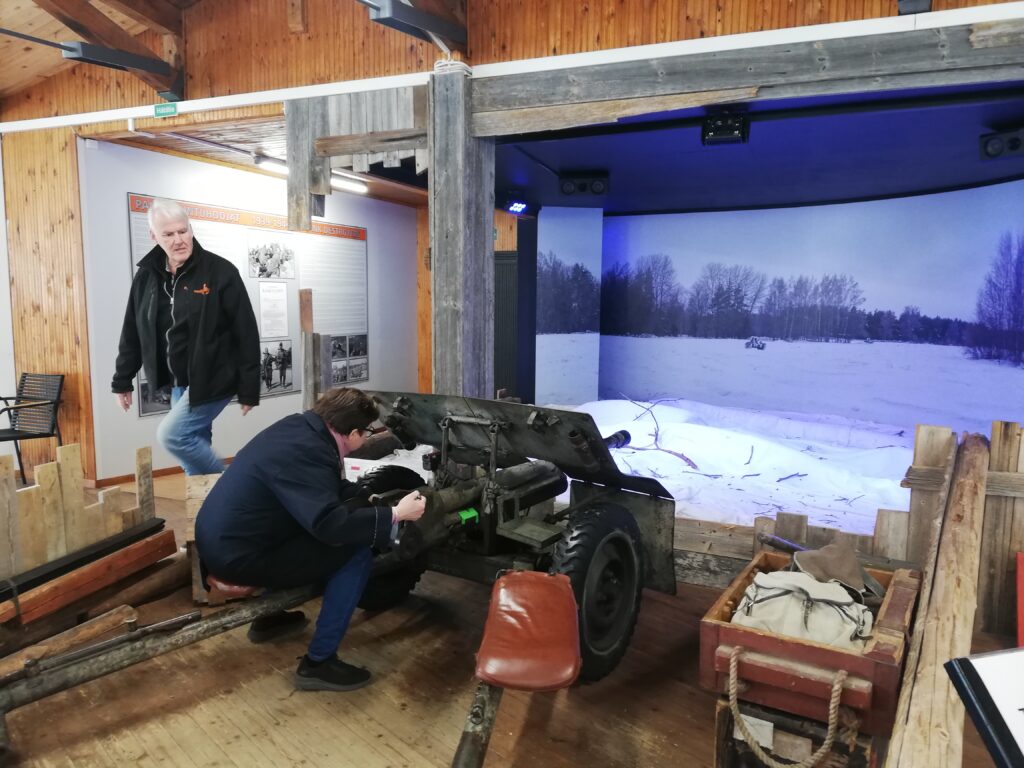 nainen istuu museossa tykin päällä. Mies kävelee taustalla. OIkealla suuri videoruutu.