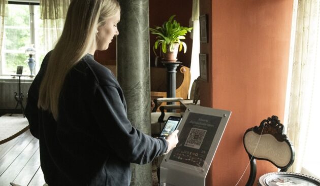 nainen tutkii museon opastaulun qr-koodia. Taustalla pylväs, viherkasvi, museon huonekaluja ja ikkuna